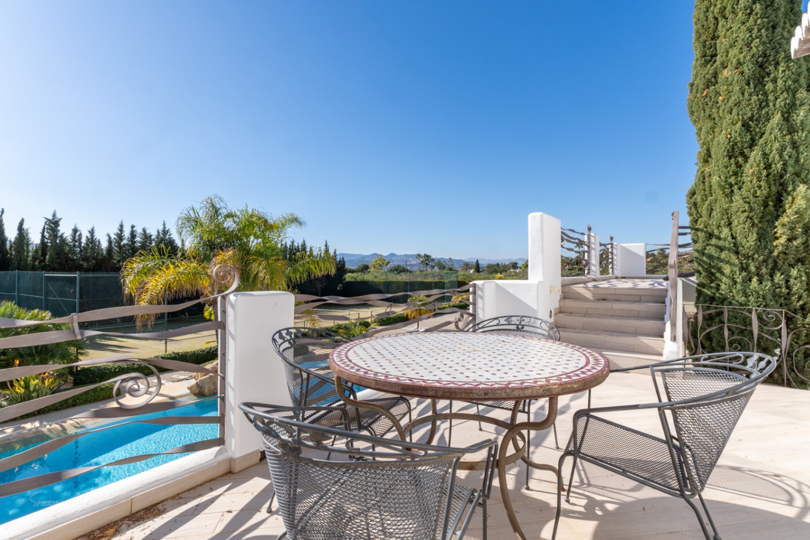 Terraza superior con mesa de comedor de mosaico y barandillas de hierro, con vistas a la piscina, la pista de tenis privada y el paisaje montañoso.