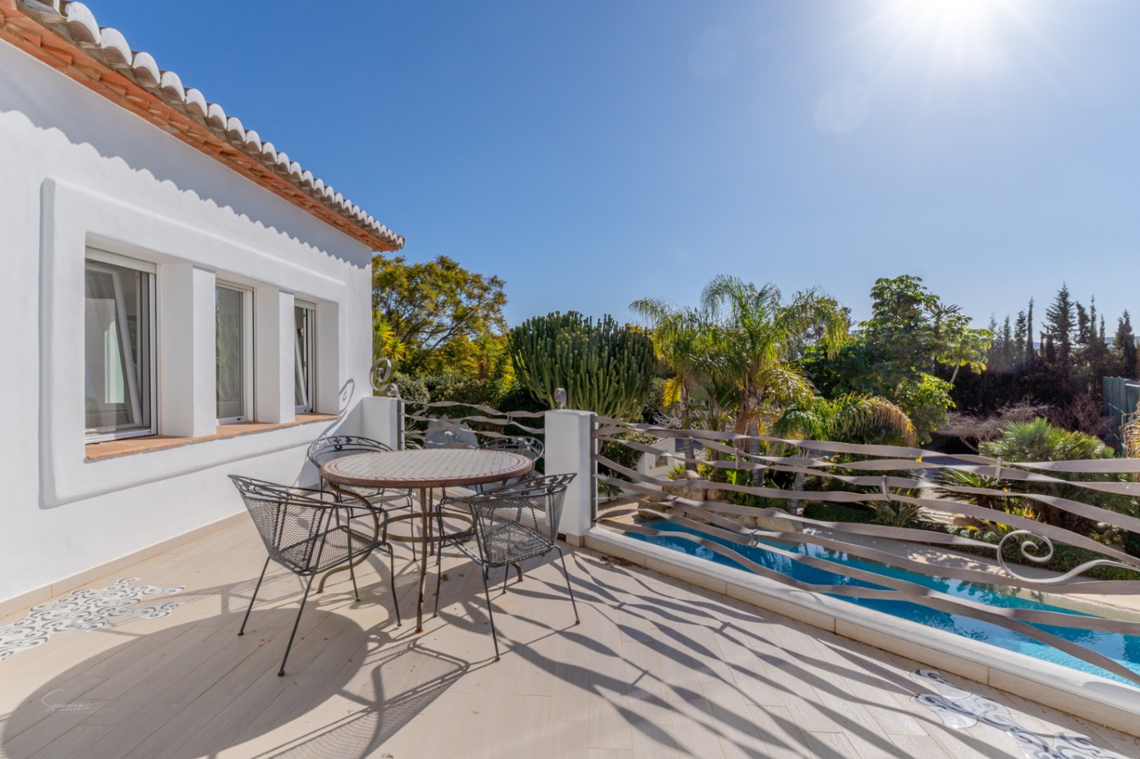 Terraza superior con suelo cerámico, mesa de mosaico y barandilla de hierro forjado ondulada, con vistas directas a la piscina y al frondoso jardín mediterráneo.