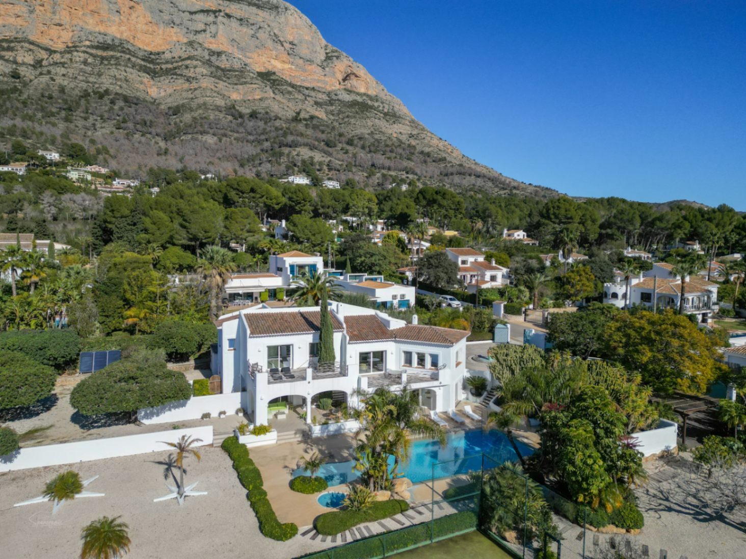 Vista aérea de la villa estilo Ibiza con fachada blanca y tejados de teja, destacando la piscina, pista de tenis y el majestuoso Montgó al fondo.