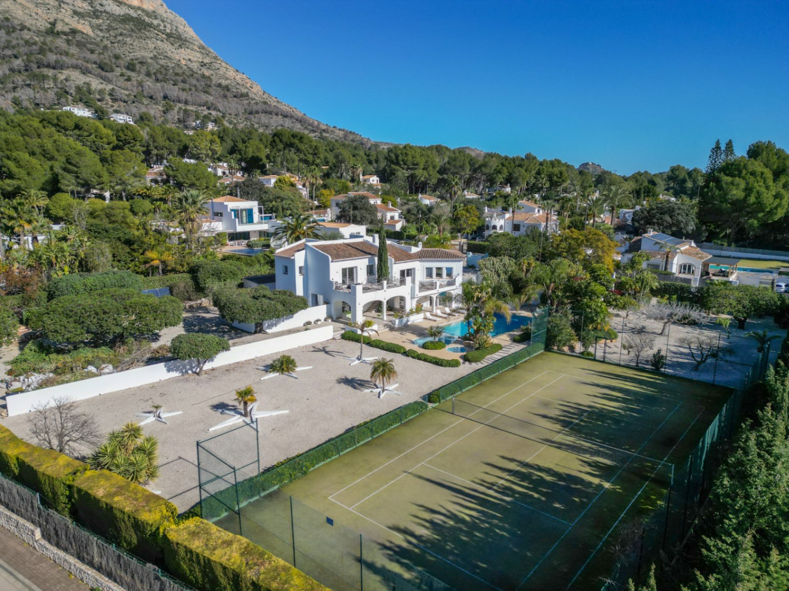 Vista aérea de la villa estilo Ibiza con pista de tenis privada, piscina rodeada de palmeras y la montaña Montgó como telón de fondo.