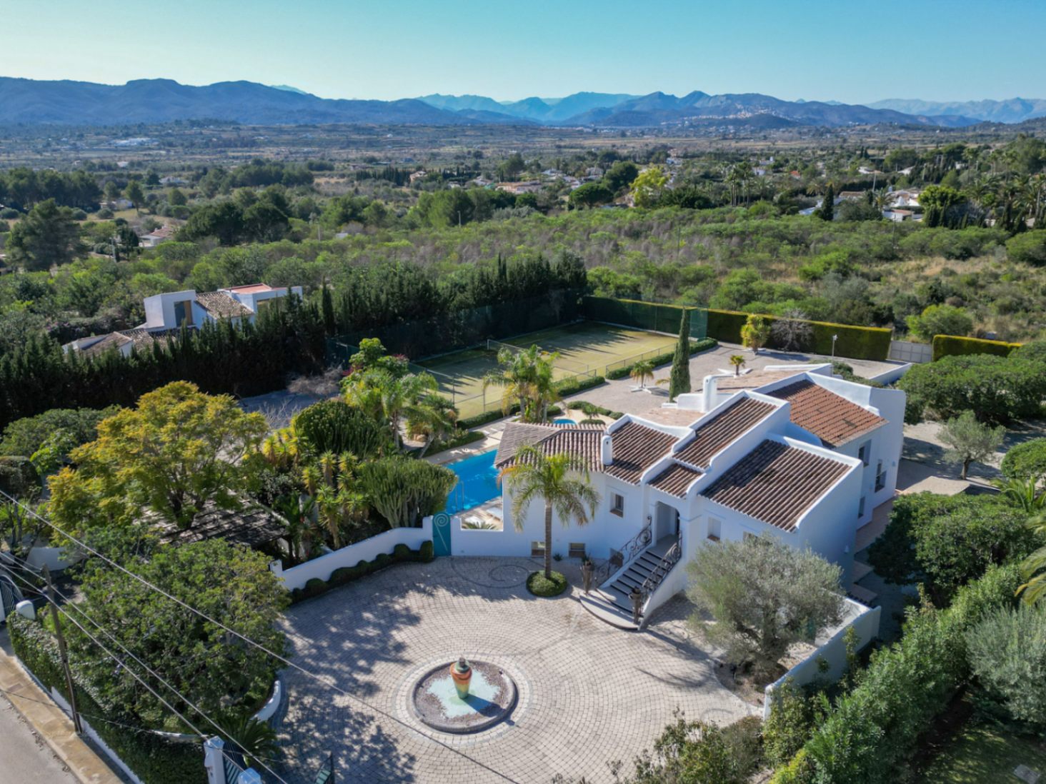 Vista aérea de la villa estilo Ibiza con su jardín pavimentado, fuente central, piscina y pista de tenis privada, enmarcada por el valle y el macizo del Montgó.