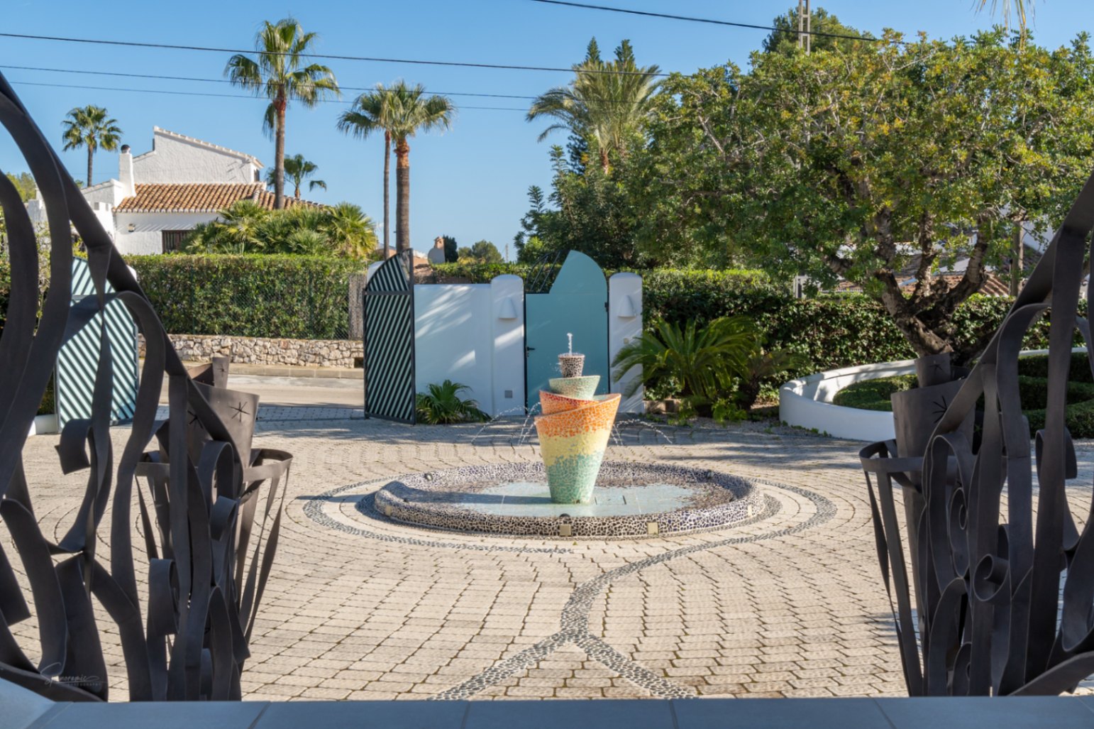 Vista desde la entrada con barandillas de hierro forjado hacia el patio pavimentado, destacando una fuente de mosaico multicolor rodeada de vegetación mediterránea.