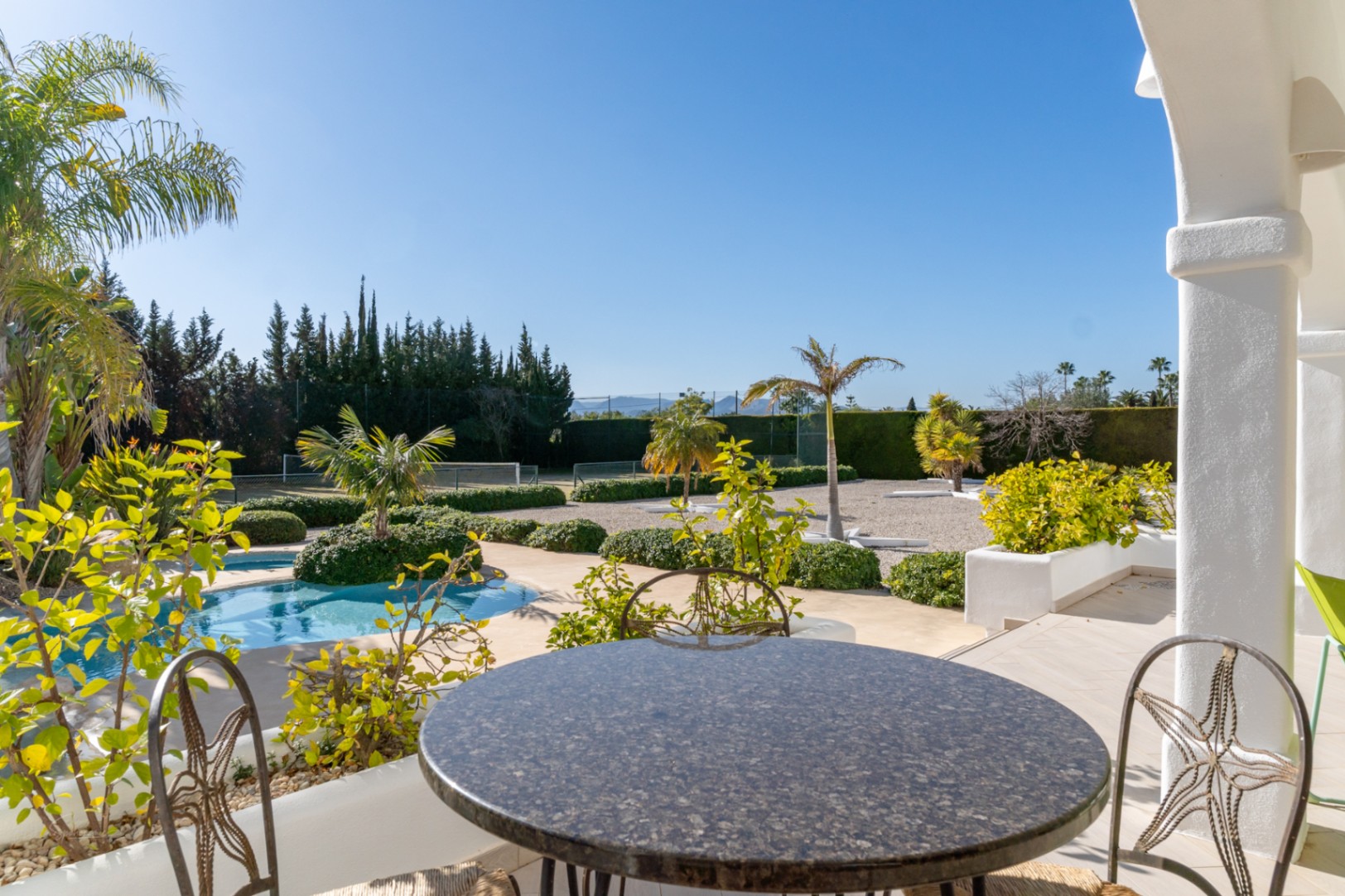 Vista desde la naya con mesa de granito, mostrando la piscina, el jardín mediterráneo y la pista de tenis privada bajo un cielo despejado.
