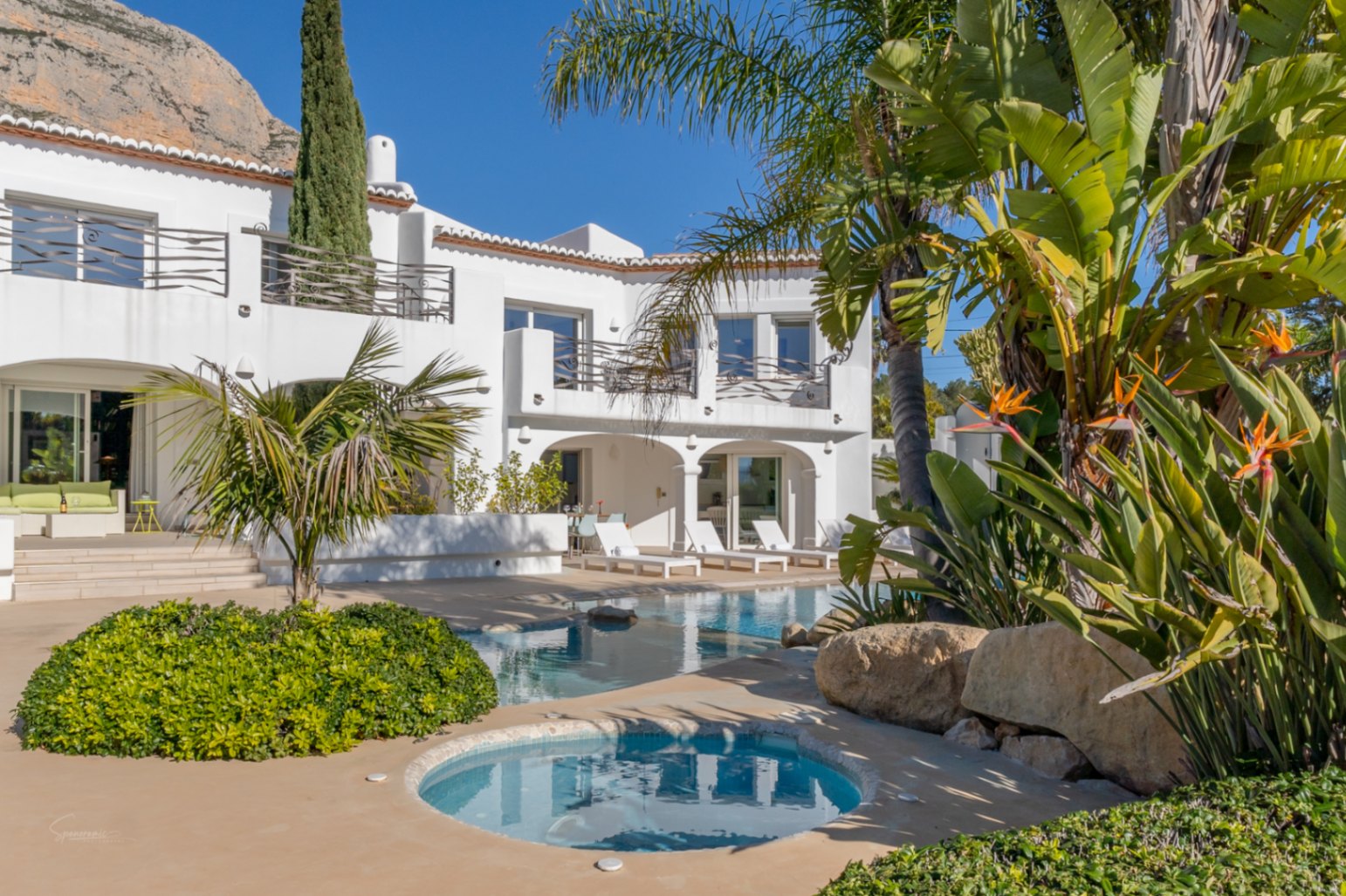 Vista exterior de la villa estilo Ibiza con piscina de diseño, jacuzzi integrado y jardines tropicales bajo el Montgó. Destaca la naya con arcos y las terrazas superiores.