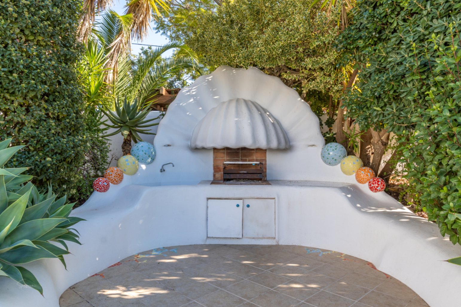 Zona de barbacoa de estilo ibicenco con estructura blanca en forma de concha, encimeras curvas, detalles de mosaico coloridos y fregadero integrado rodeado de vegetación mediterránea.