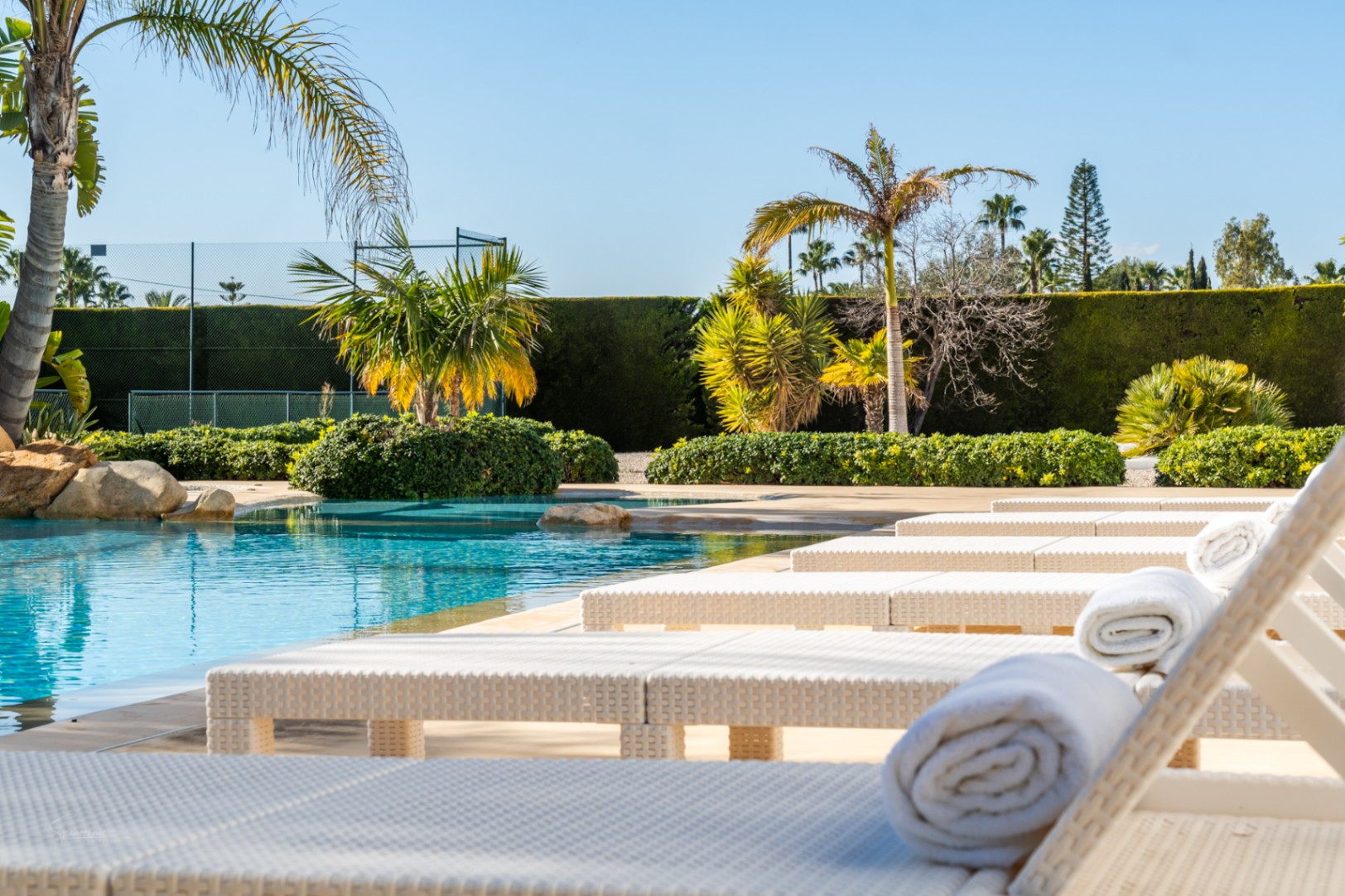 Zona de descanso junto a la piscina con tumbonas de ratán blanco y toallas, rodeada de vegetación mediterránea y una pista de tenis privada.