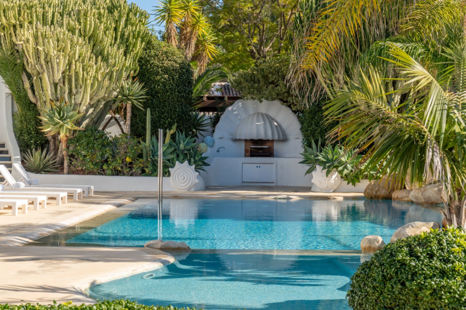 Zona de piscina con barbacoa de diseño mediterráneo en forma de concha, rodeada de vegetación exuberante, cactus y tumbonas blancas.