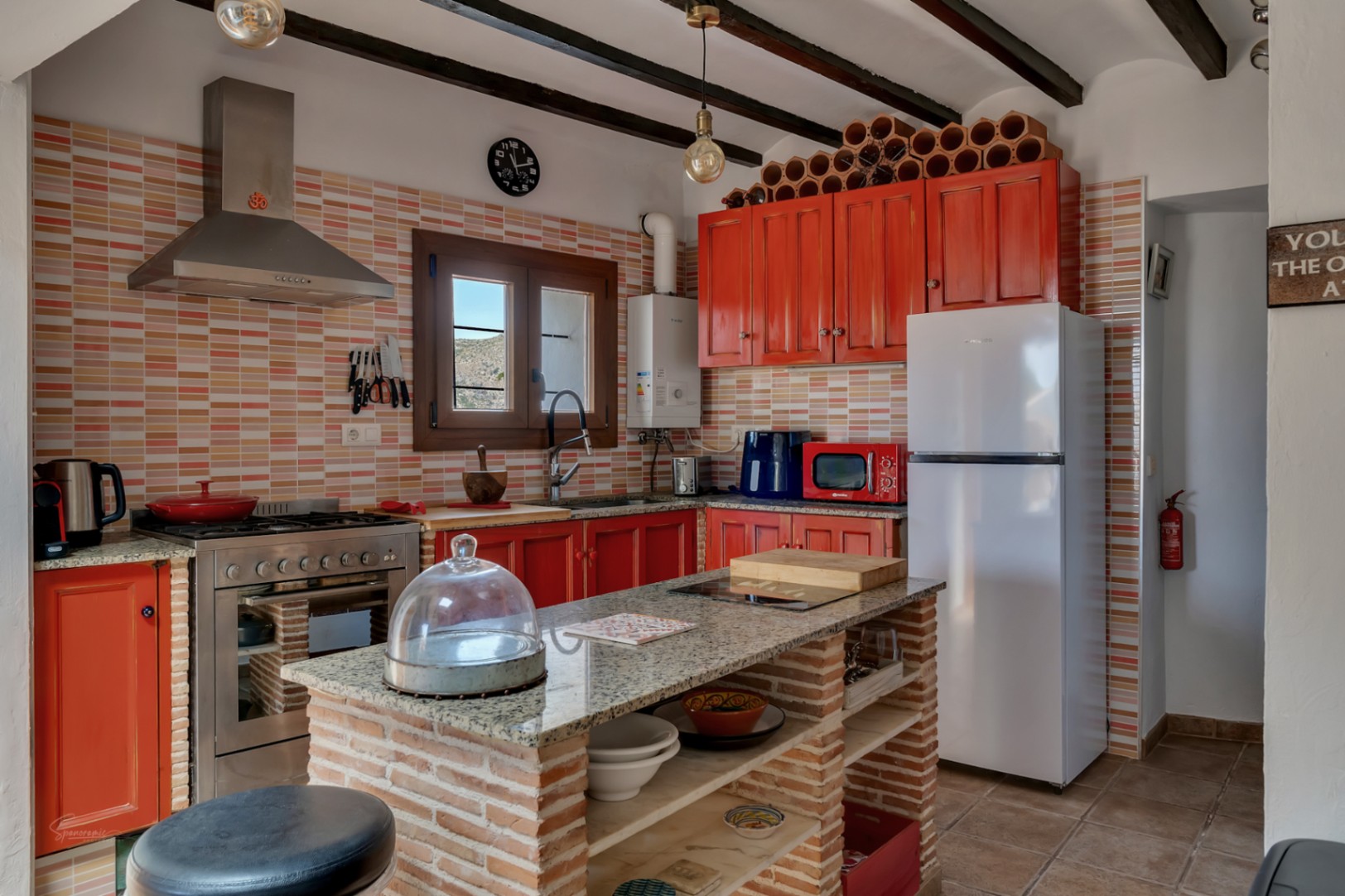 Cocina rústica con encimeras de granito, armarios rojos, y una isla central de ladrillo con estantes abiertos.