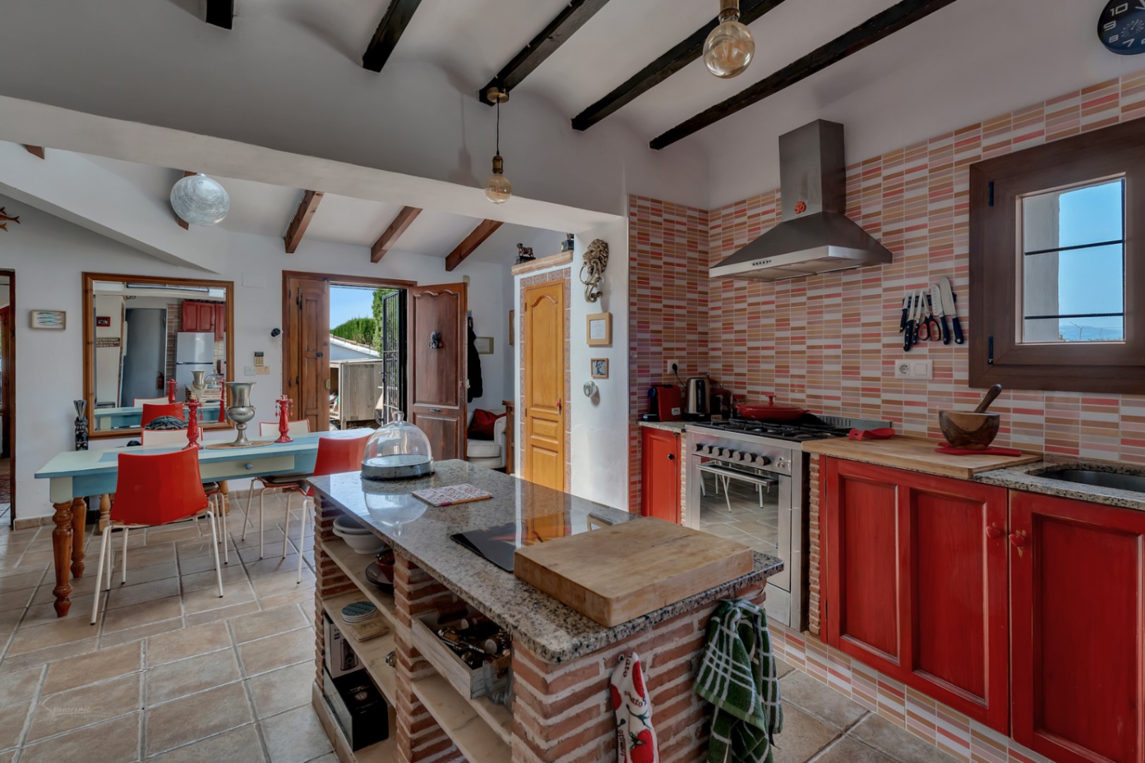 Cocina rústica con isla de ladrillo, azulejos color melocotón y muebles rojos. Vigas de madera en el techo.