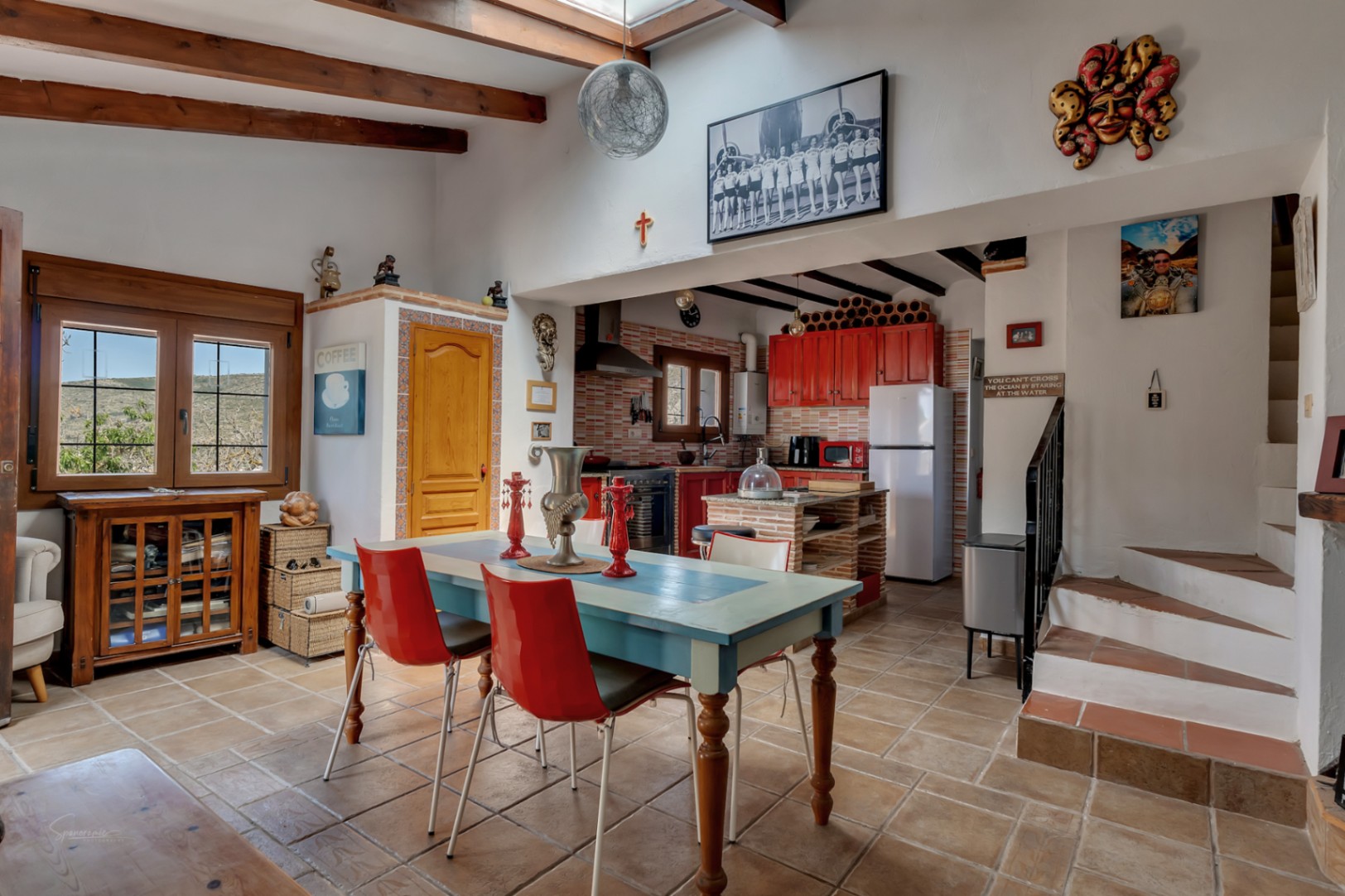 Comedor luminoso con mesa azul, sillas rojas y cocina roja. Vigas de madera y decoración ecléctica.