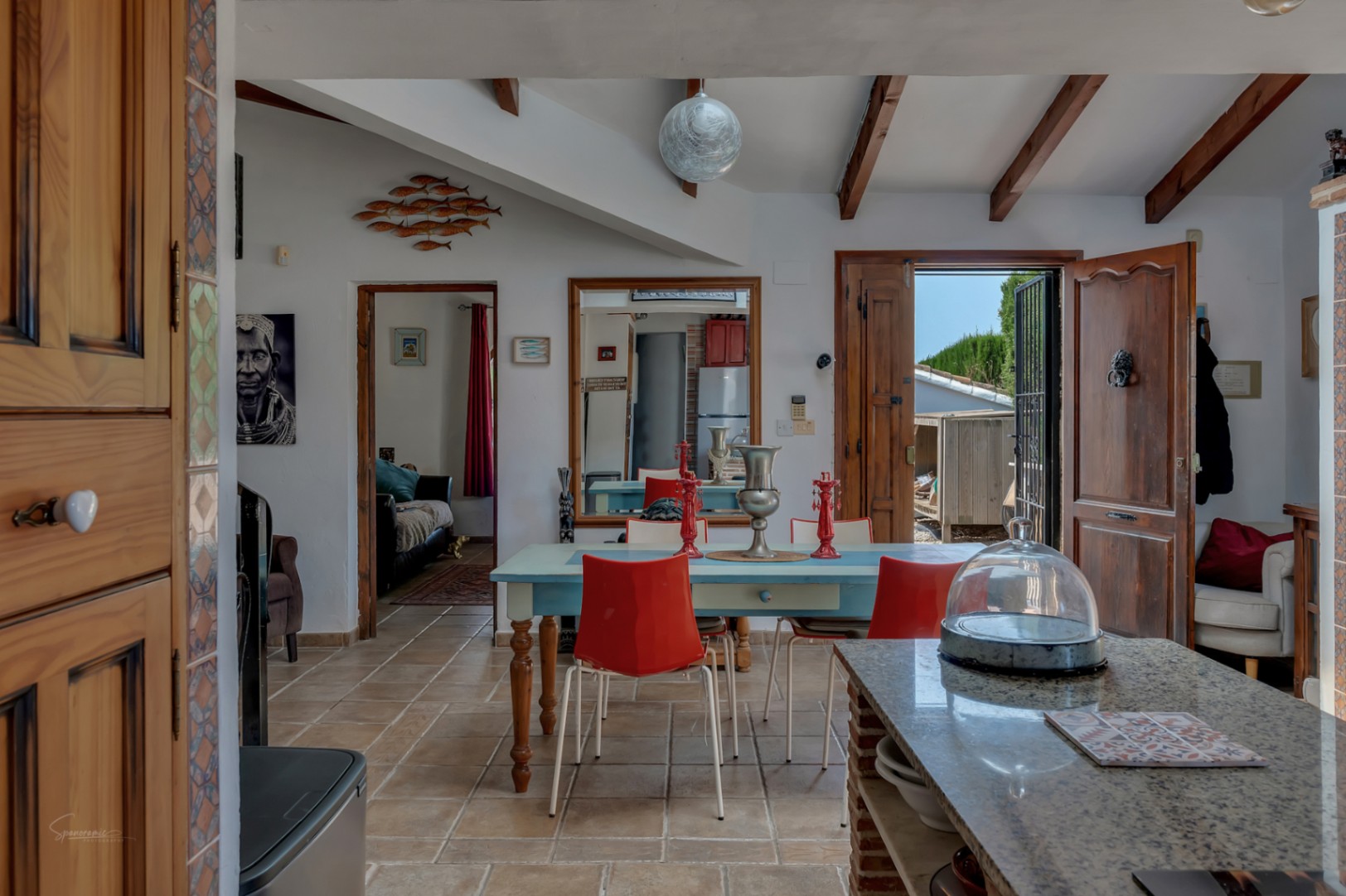 Comedor rústico con mesa azul y sillas rojas, suelo de baldosas y vigas de madera.