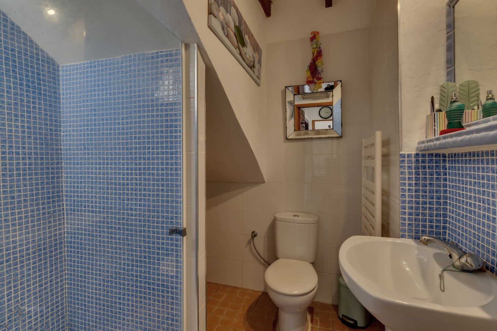 Detalle de baño con ducha de mosaico azul, inodoro blanco y lavabo con encimera de mosaico.