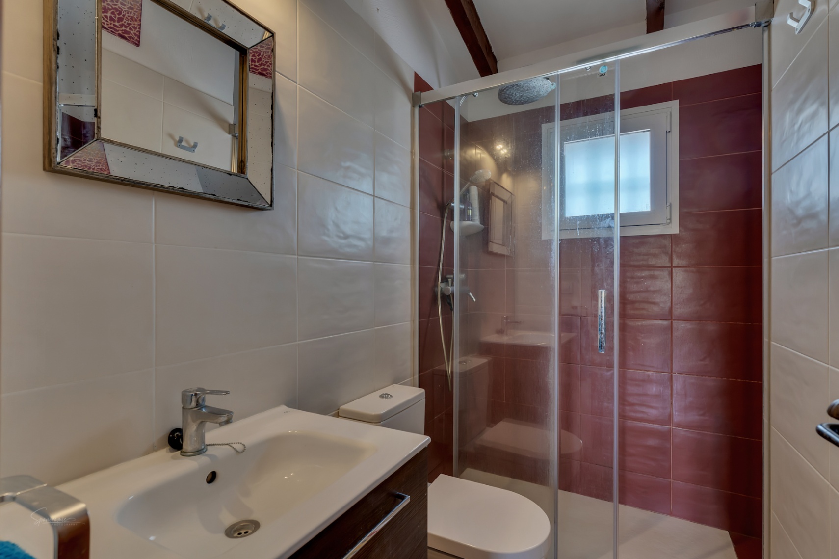 Detalle de baño con ducha de vidrio, lavabo moderno y azulejos en tonos neutros y burdeos.