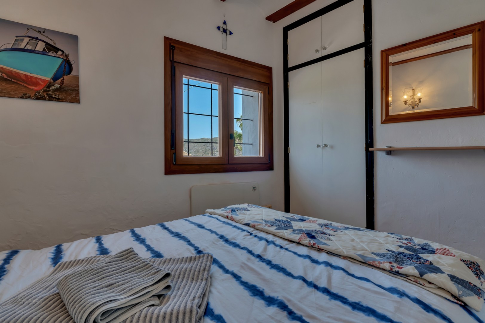 Dormitorio con ventana de madera, armario blanco y ropa de cama a rayas azules. Decoración rústica y ambiente luminoso.