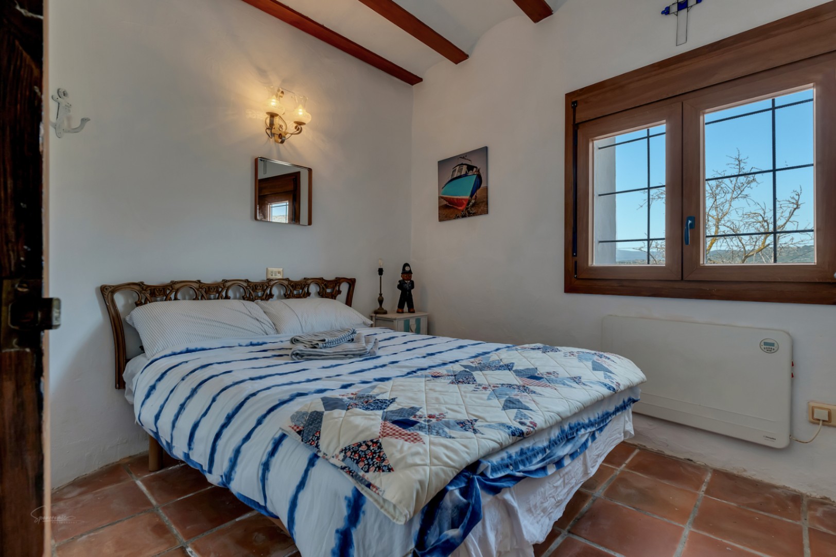 Dormitorio rústico con suelo de terracota, cama con colcha azul y ventana de madera con vistas al exterior.