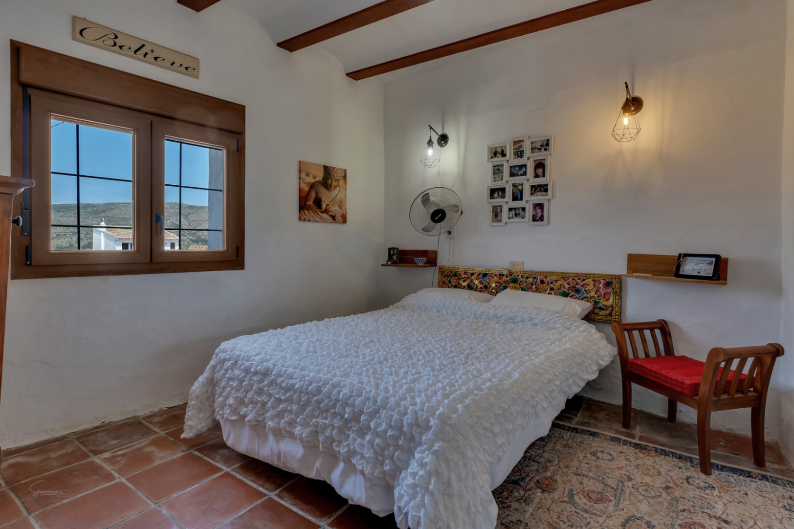 Habitación rústica con suelo de terracota, cama doble con cabecero tallado y ventana con vistas al exterior.