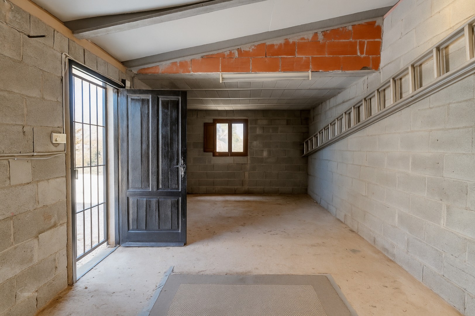 Interior de garaje con paredes de bloques de hormigón, puerta de madera abierta y escalera metálica.