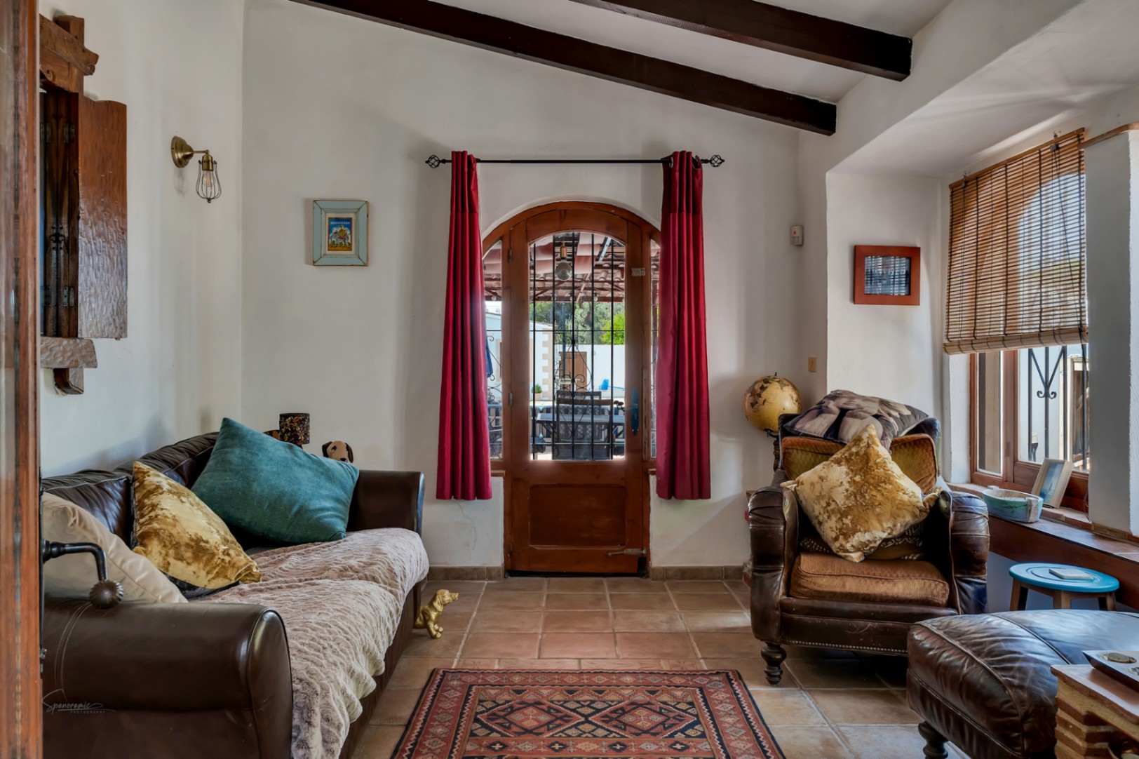 Salón rústico con sofá de cuero, sillón vintage y puerta arqueada con cortinas rojas en Teulada.