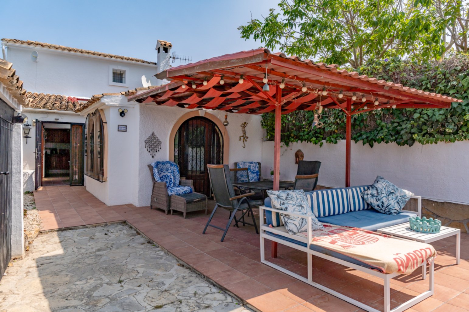Terraza cubierta con toldo rojo, mobiliario de jardín y acceso a la casa de campo en Teulada.