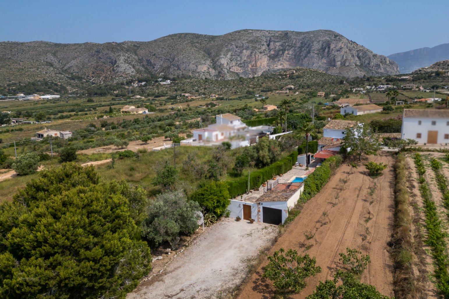 Vista aérea de la propiedad rural con camino de tierra, garaje y piscina. Campos cultivados y montañas rocosas en el horizonte definen el entorno natural de Teulada.