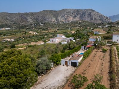 Vista aérea de la propiedad rural con camino de tierra, garaje y piscina. Campos cultivados y montañas rocosas en el horizonte definen el entorno natural de Teulada.