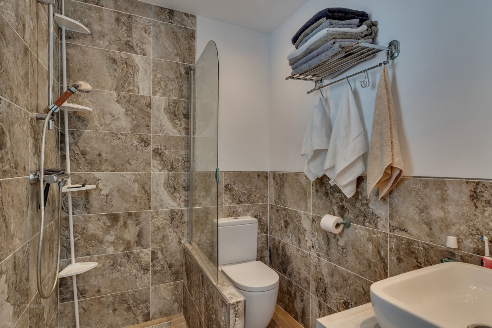 Detalle de baño con ducha, inodoro y lavabo. Azulejos marrones veteados y toallas.