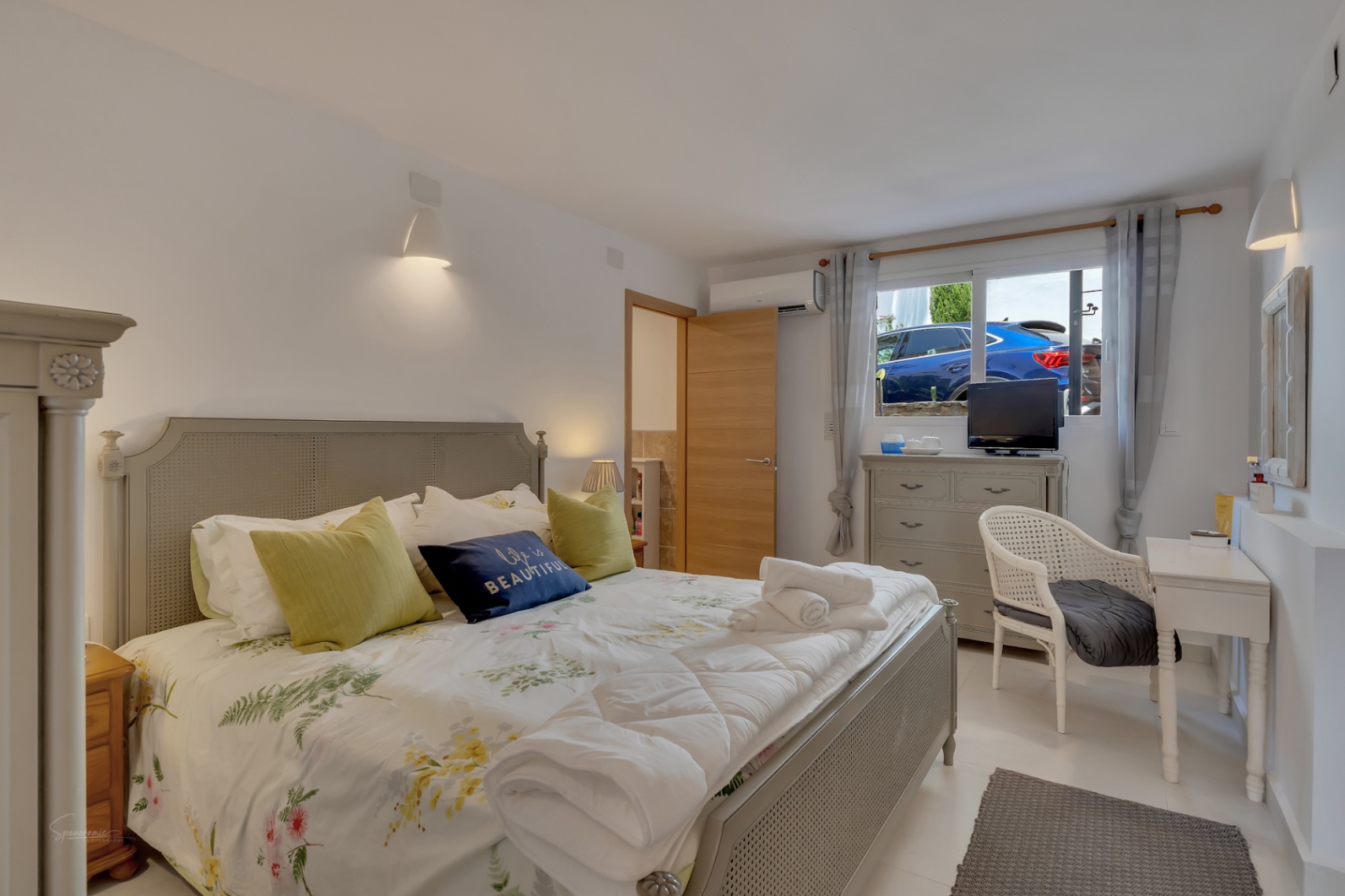 Dormitorio luminoso con cama de rejilla gris, ropa de cama floral y tocador blanco con silla de mimbre.