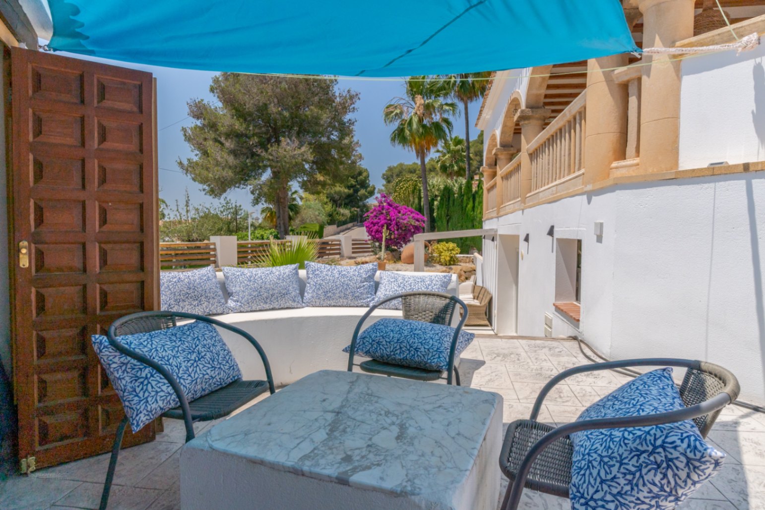 Terraza con asientos de obra, cojines estampados y mesa de mármol, bajo toldo azul.