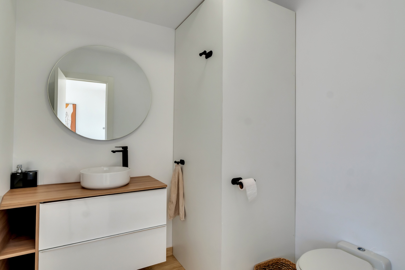 Aseo de diseño moderno con lavabo sobre encimera de madera, grifería negra mate y espejo circular. El mueble suspendido blanco aporta un estilo minimalista y funcional.