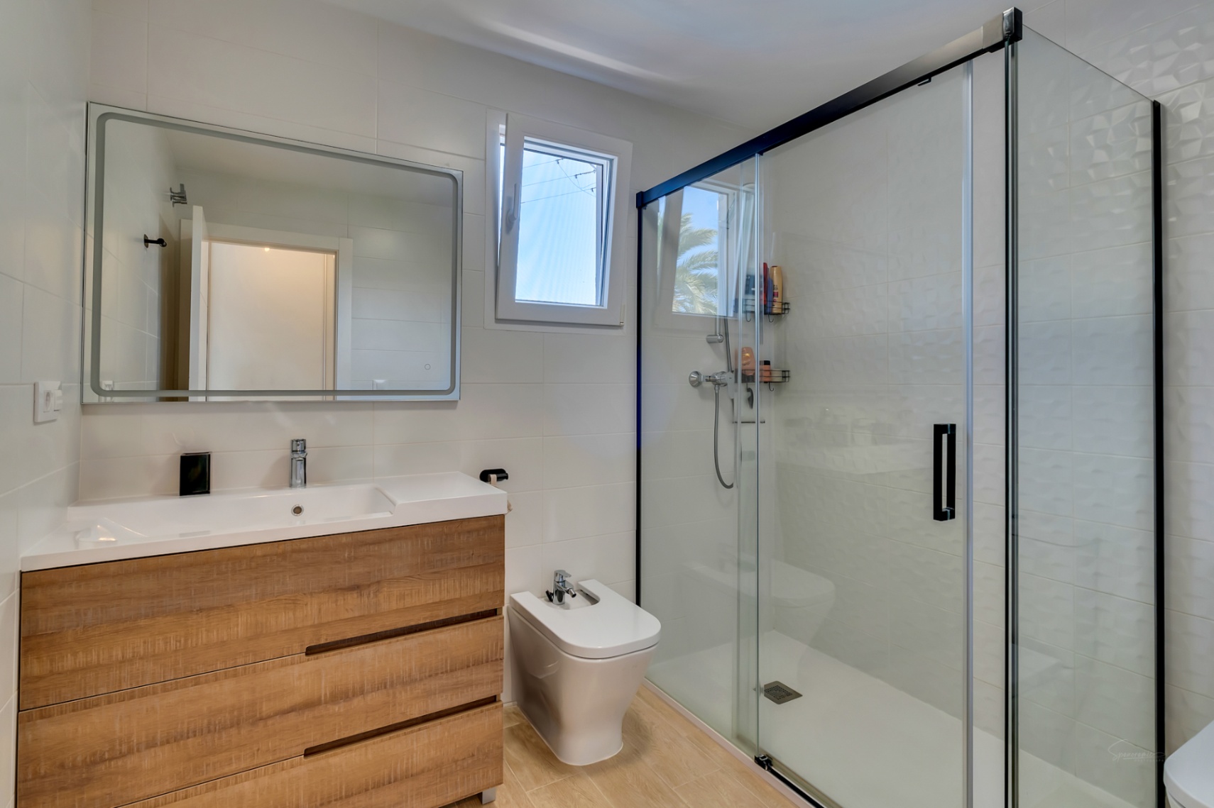 Baño contemporáneo con mueble de lavabo efecto madera, bidé y una amplia ducha con mampara de cristal y perfilería negra. Incluye ventana para ventilación natural.