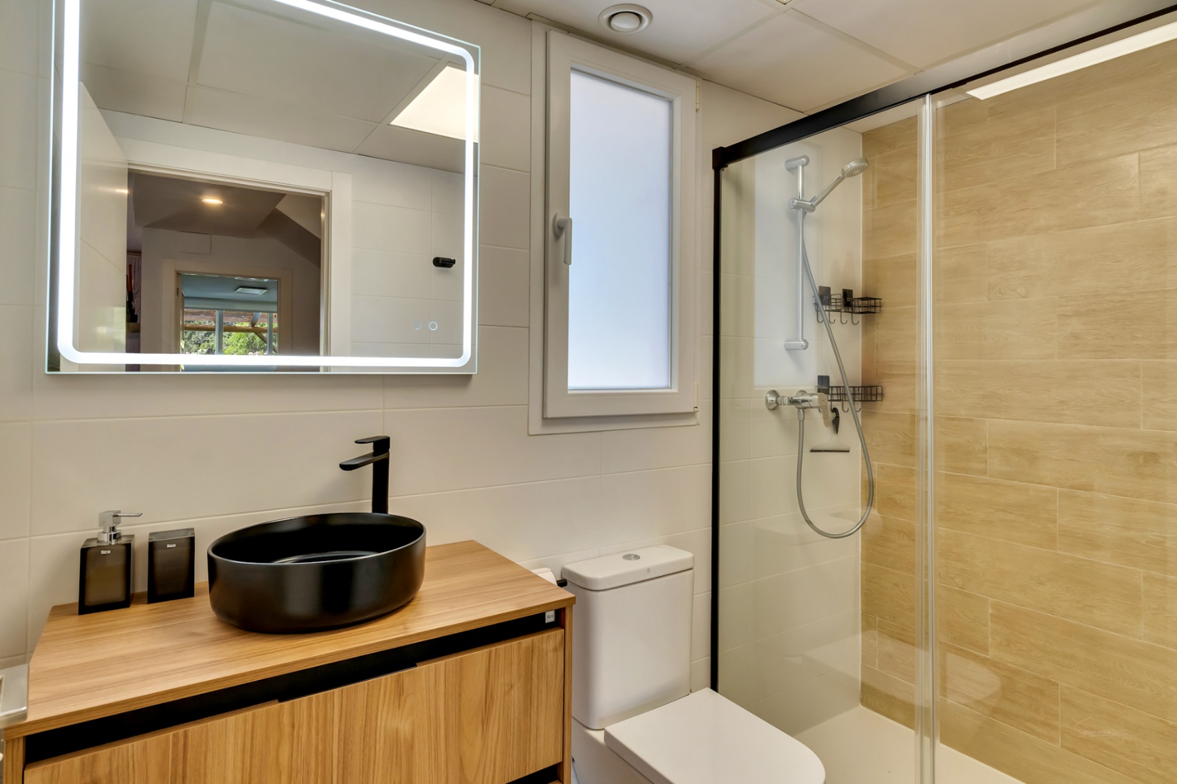 Baño moderno con lavabo sobre encimera negro, mueble de madera y espejo retroiluminado. Incluye ducha con mampara de perfil negro y azulejos efecto madera.