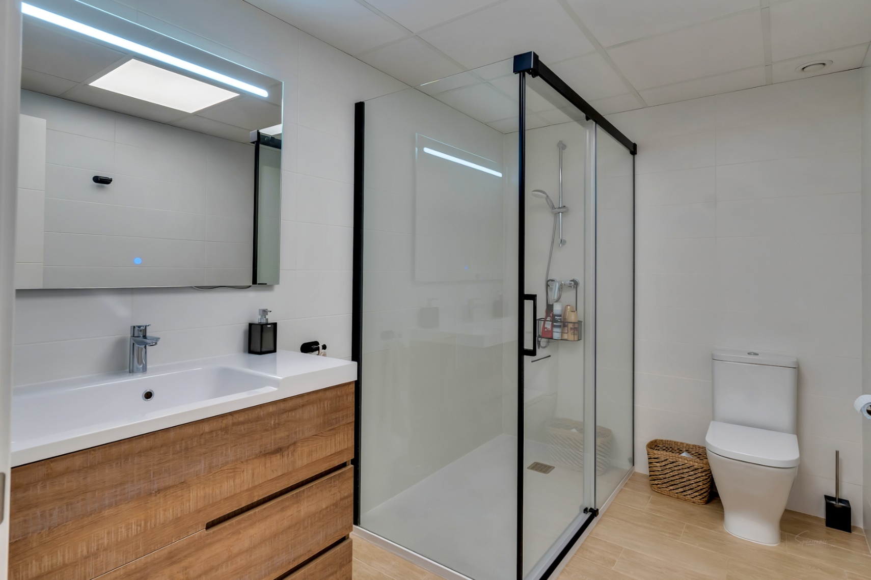 Baño moderno con mueble de madera, espejo con iluminación LED integrada y amplia ducha con mampara de perfilería negra y suelo de gres efecto madera.