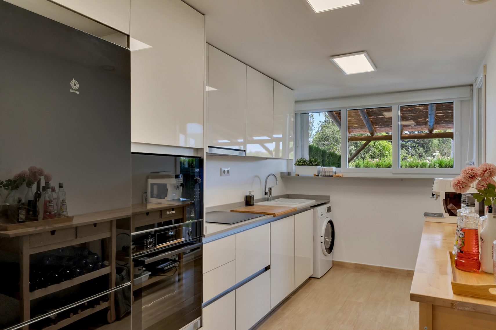 Cocina moderna con armarios blancos de alto brillo, electrodomésticos integrados y ventanal con vistas al patio privado y la pérgola de madera.