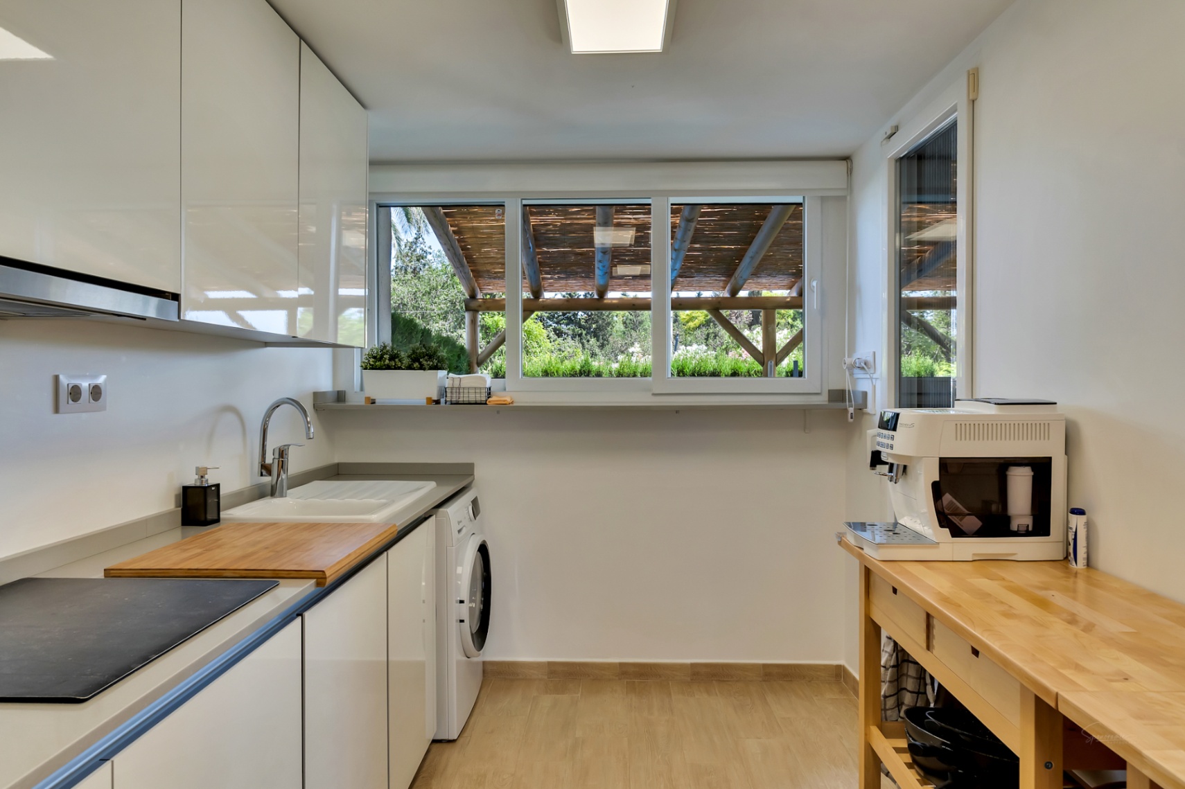 Cocina moderna con armarios blancos, zona de lavandería integrada y mesa auxiliar de madera. El amplio ventanal aporta luz natural y vistas a la pérgola del patio.