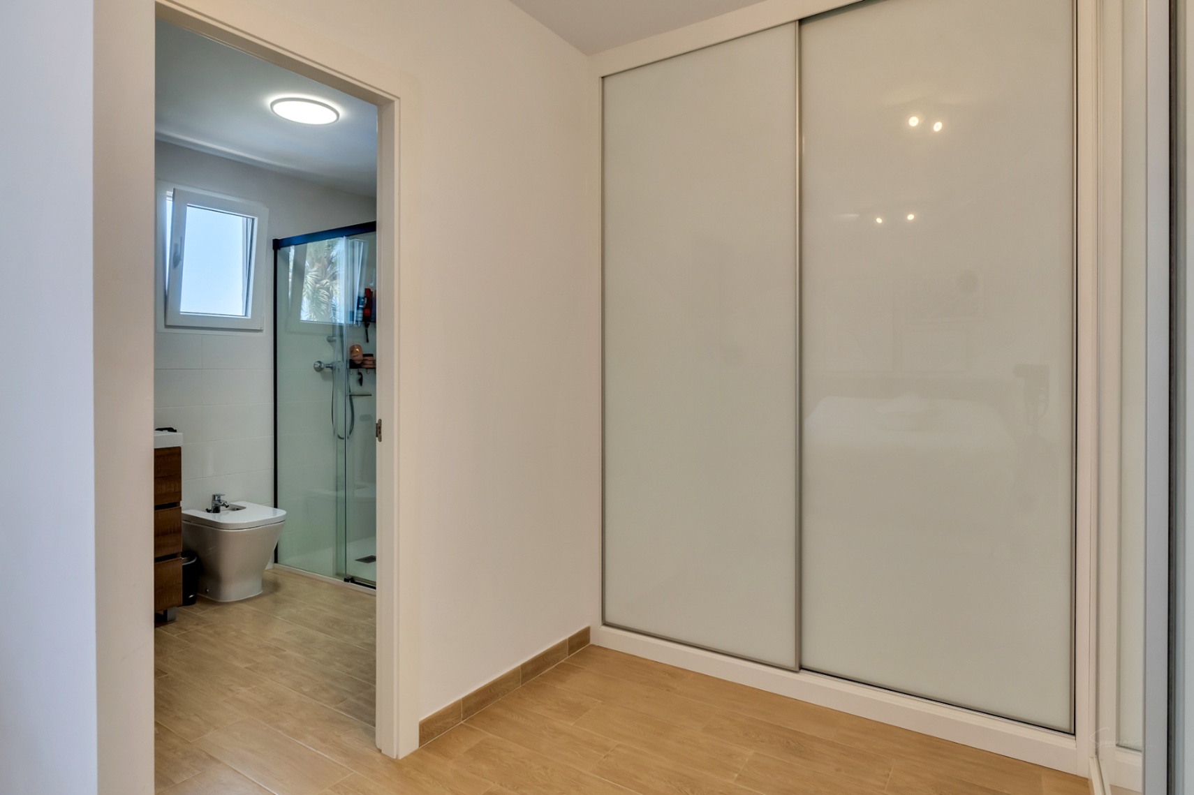 Dormitorio con amplio armario empotrado de puertas correderas blancas y acceso directo al baño en suite con ducha y acabados modernos.