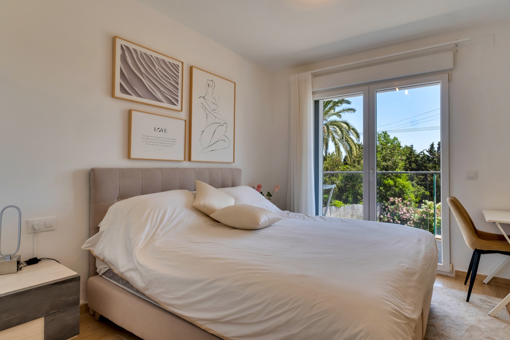 Dormitorio doble con cabecero tapizado, zona de escritorio y acceso a un balcón con vistas despejadas a la vegetación mediterránea.