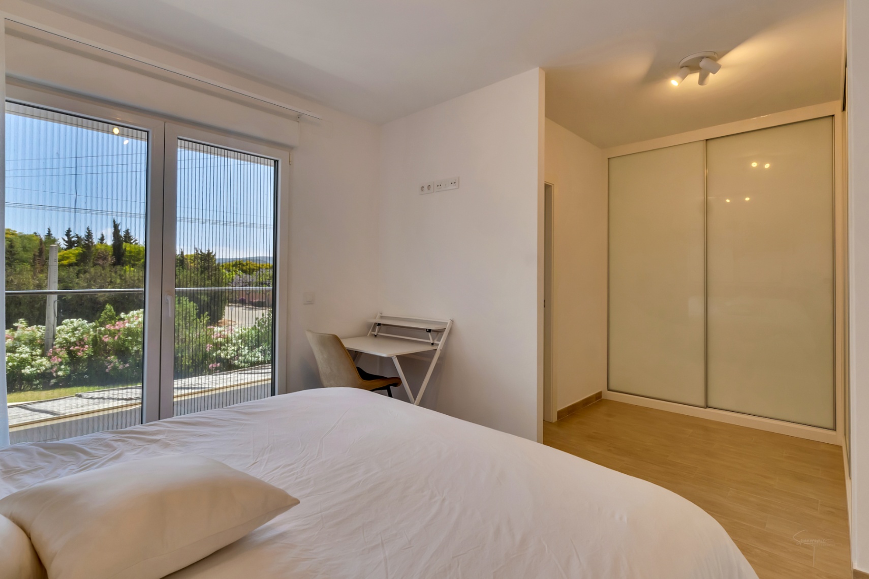 Dormitorio luminoso con armario empotrado de puertas correderas blancas, zona de escritorio y acceso directo a una terraza con vistas despejadas y mosquitera integrada.