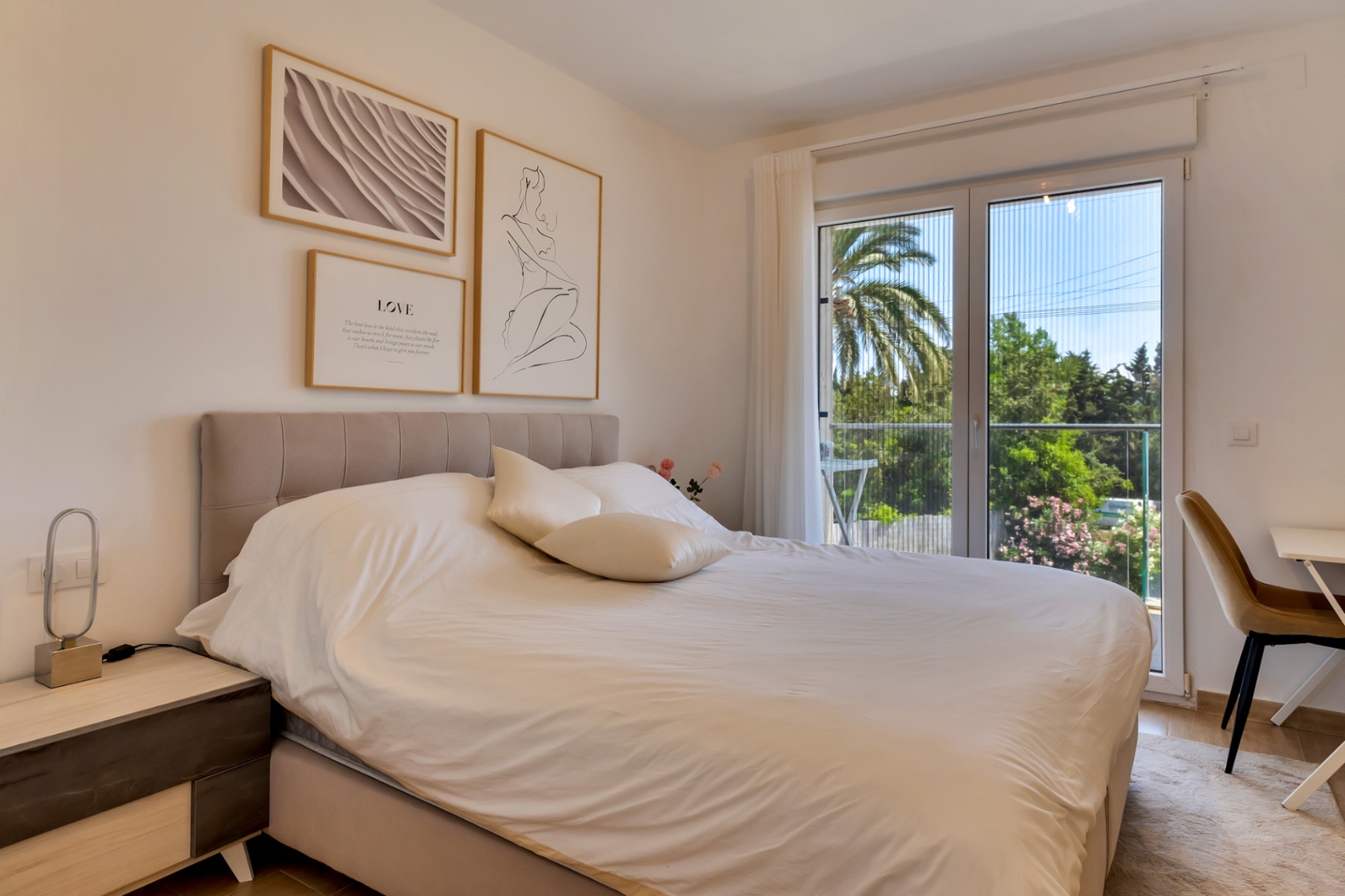 Dormitorio luminoso con cama doble, cabecero tapizado y zona de escritorio. Una gran puerta acristalada con mosquitera da acceso al balcón con vistas a la vegetación.