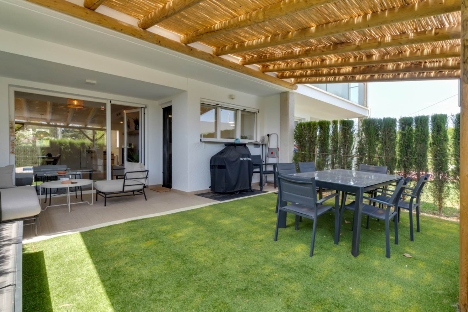 Patio privado con césped artificial, pérgola de madera y zona de comedor exterior. Dispone de área de estar y barbacoa, conectado al interior por amplios ventanales.