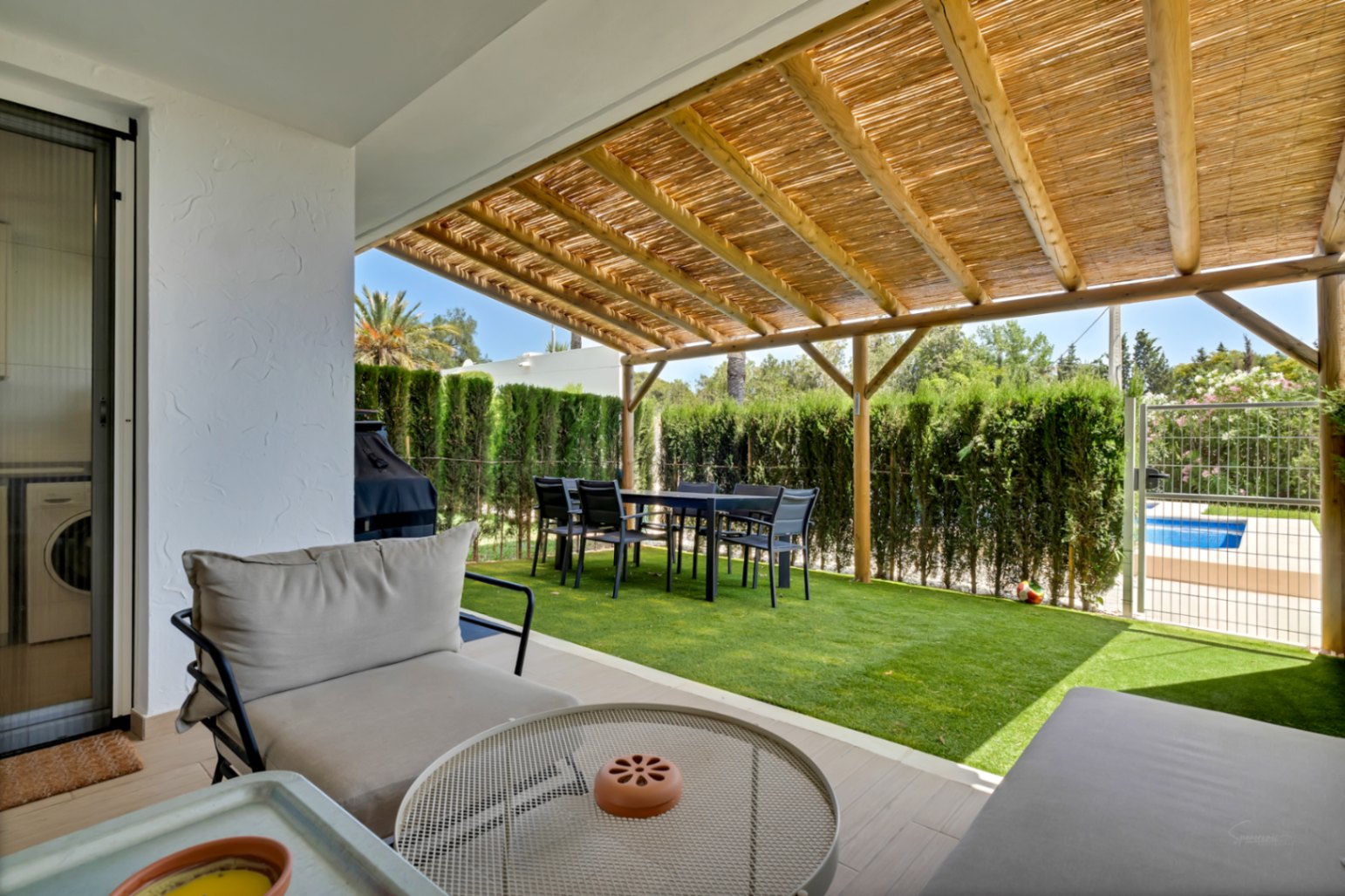 Patio privado con pérgola de madera y cañizo, zona de comedor exterior y césped artificial. Incluye acceso directo a la piscina comunitaria a través de una puerta privada.