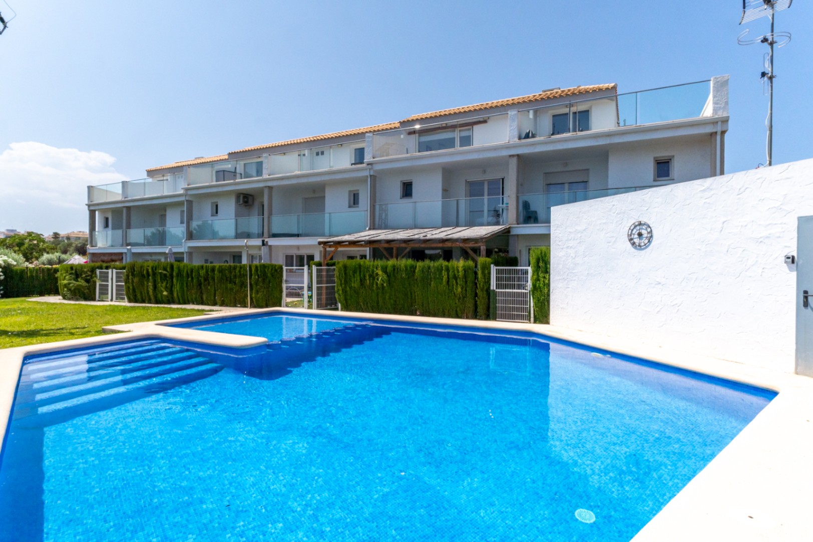 Piscina comunitaria de gresite azul frente a la fachada moderna de las viviendas, destacando sus barandillas de cristal y patios privados delimitados por setos.