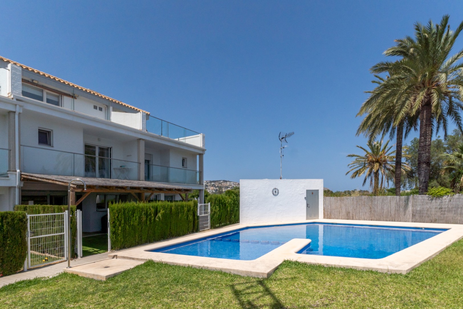 Piscina comunitaria rodeada de césped y palmeras, con acceso directo desde el patio privado. Se aprecia la fachada moderna con barandillas de cristal y pérgola de madera.