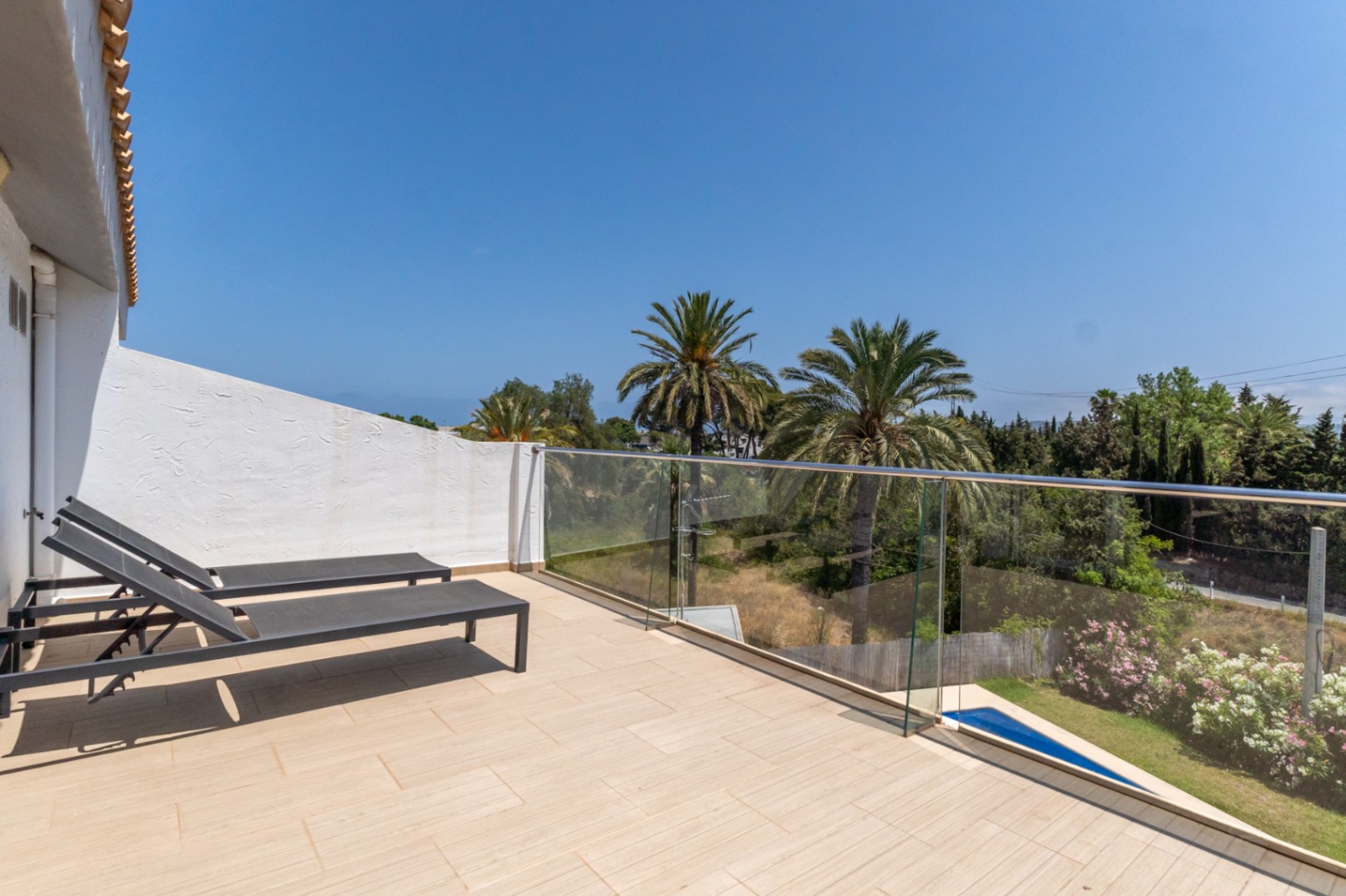 Terraza superior con suelo cerámico, barandilla de cristal y tumbonas, ofreciendo vistas despejadas a las palmeras y zonas verdes circundantes.
