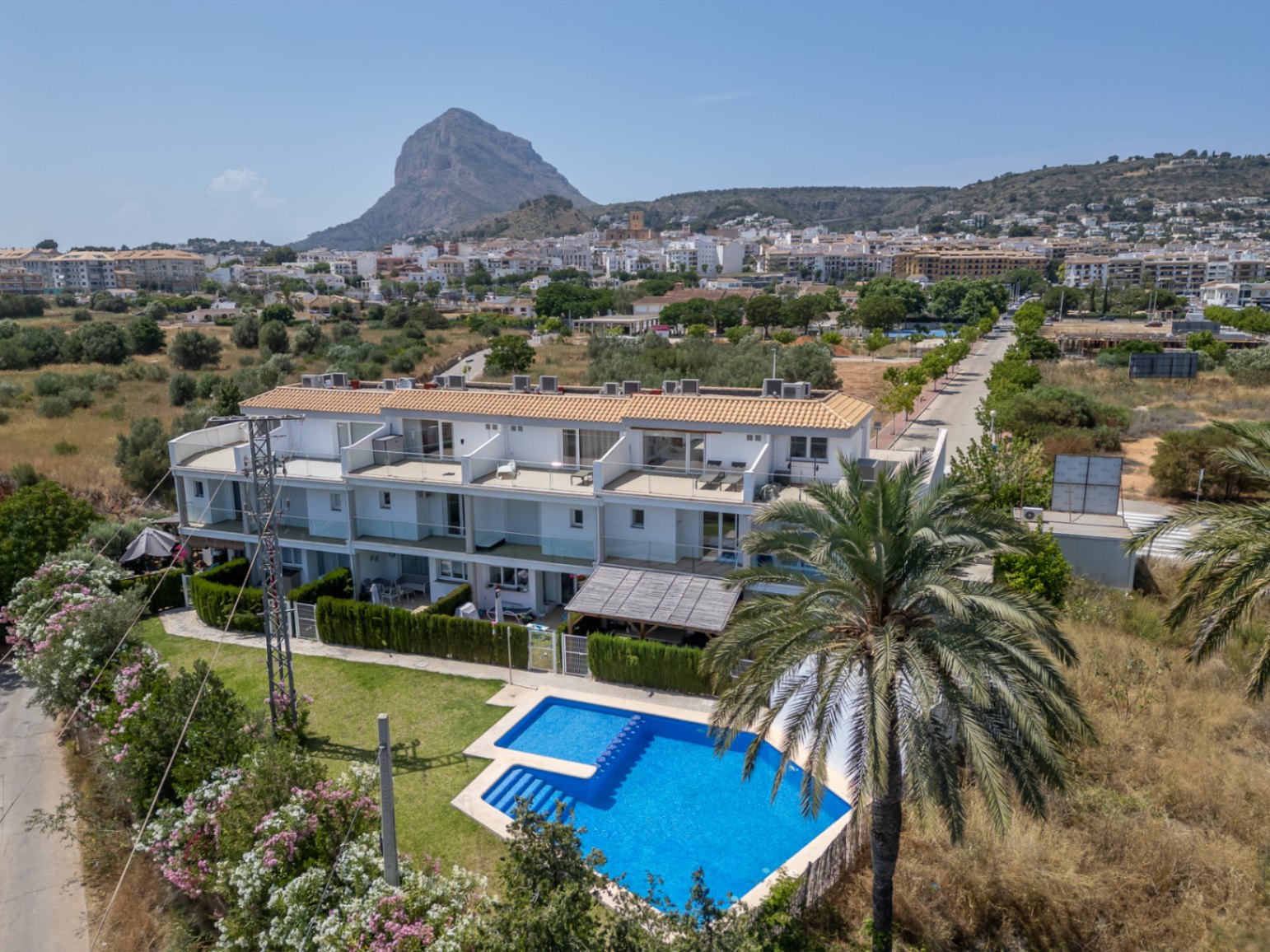 Vista aérea del complejo residencial con piscina comunitaria y jardines, bajo la silueta del Montgó. Destacan las fachadas blancas modernas y las amplias terrazas superiores.