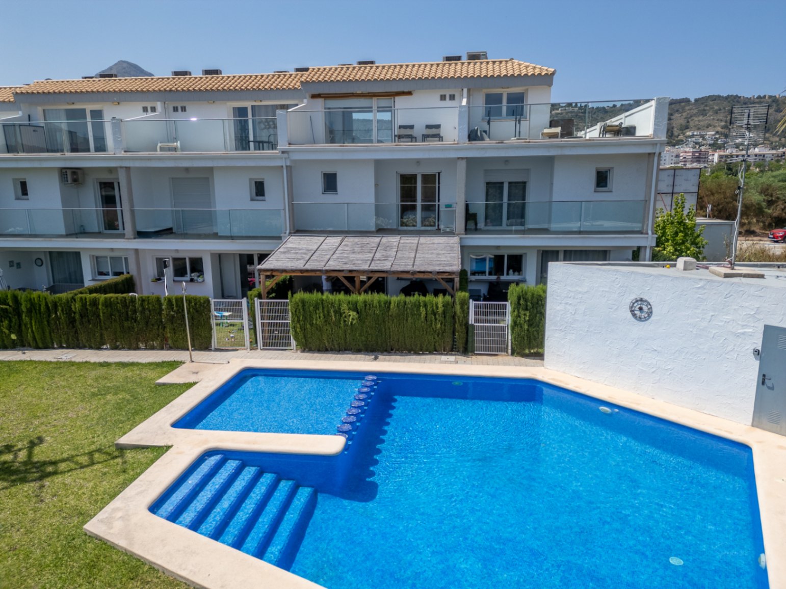 Vista de la piscina comunitaria y la fachada sur de la vivienda, con su patio privado bajo pérgola de madera y amplias terrazas con barandillas de cristal.