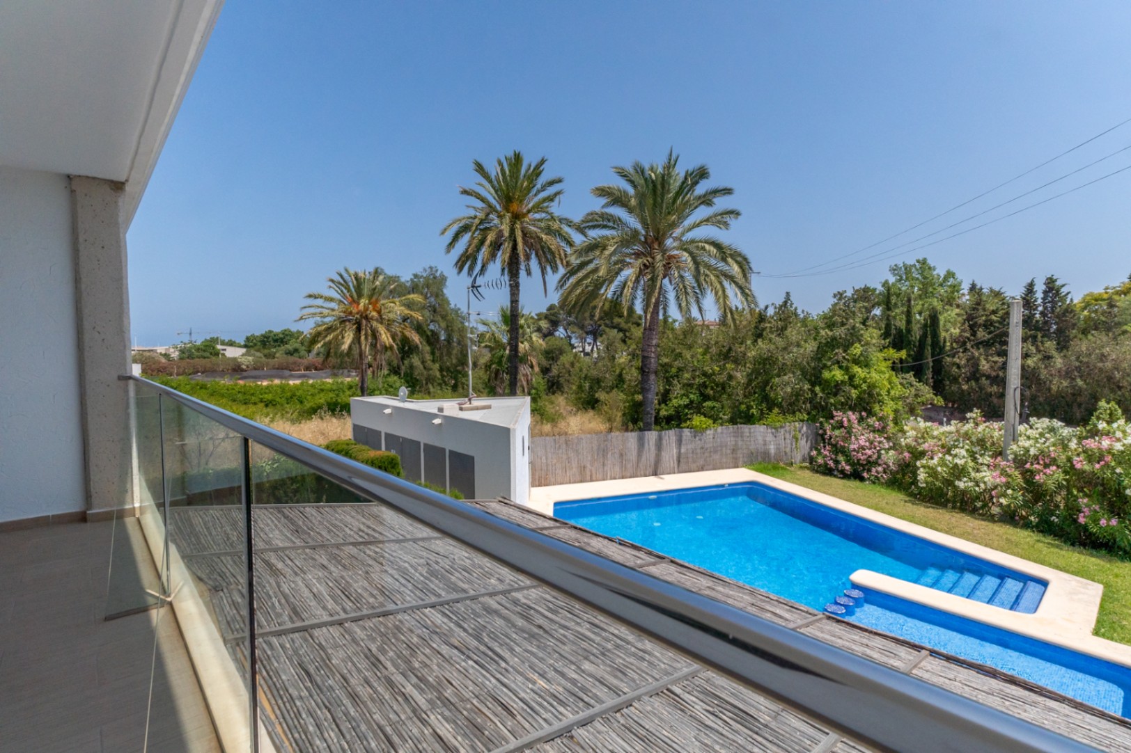 Vista desde la terraza superior con barandilla de cristal, orientada a la piscina comunitaria y rodeada de palmeras y vegetación mediterránea.