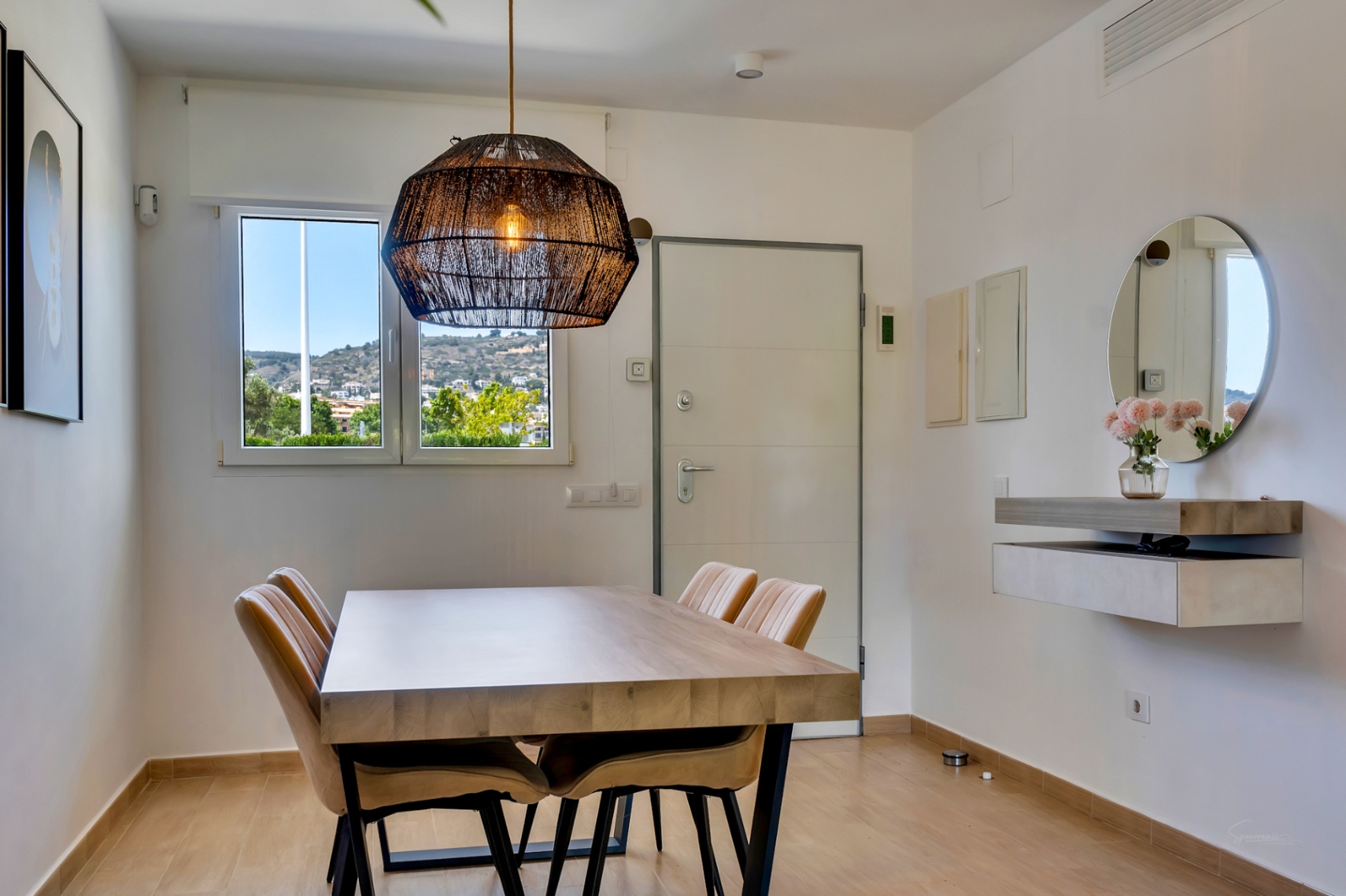 Zona de comedor con mesa de madera maciza, sillas tapizadas y lámpara de fibras naturales. Incluye un mueble flotante con espejo circular y ventana con vistas al entorno.