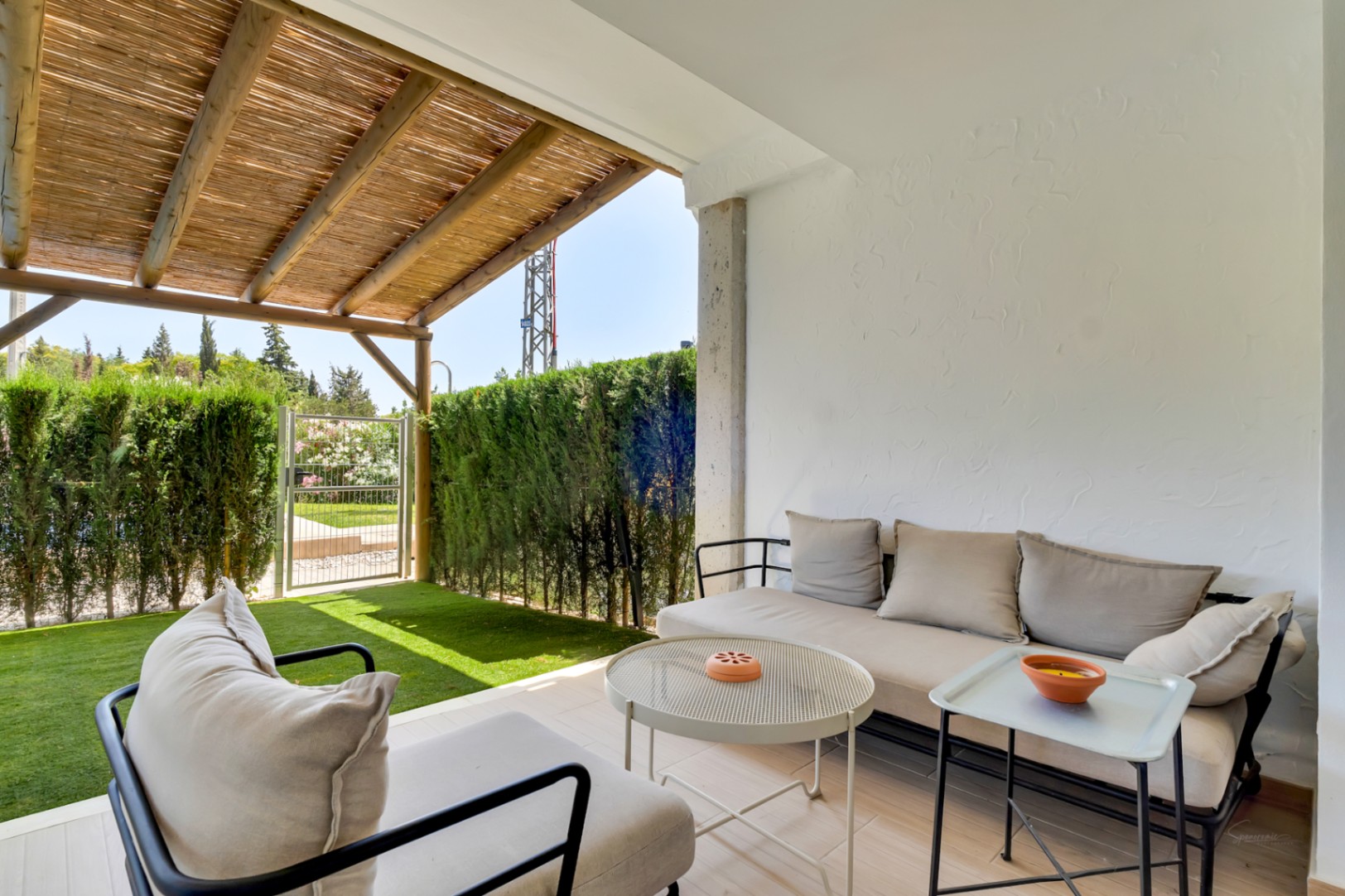 Zona de estar exterior en el patio privado, protegida por una pérgola de madera y cañizo, con mobiliario moderno y acceso directo al jardín de césped artificial y setos.