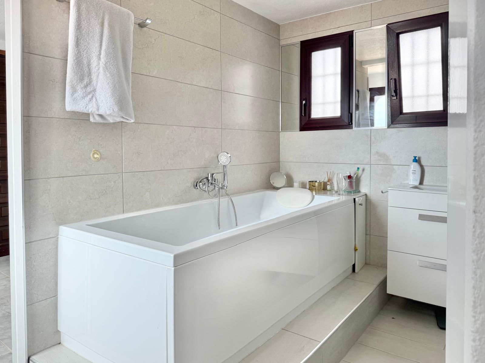 Baño con bañera blanca, paredes de gres en tonos neutros y mueble de lavabo funcional. Destaca la carpintería oscura de las ventanas y la grifería cromada.