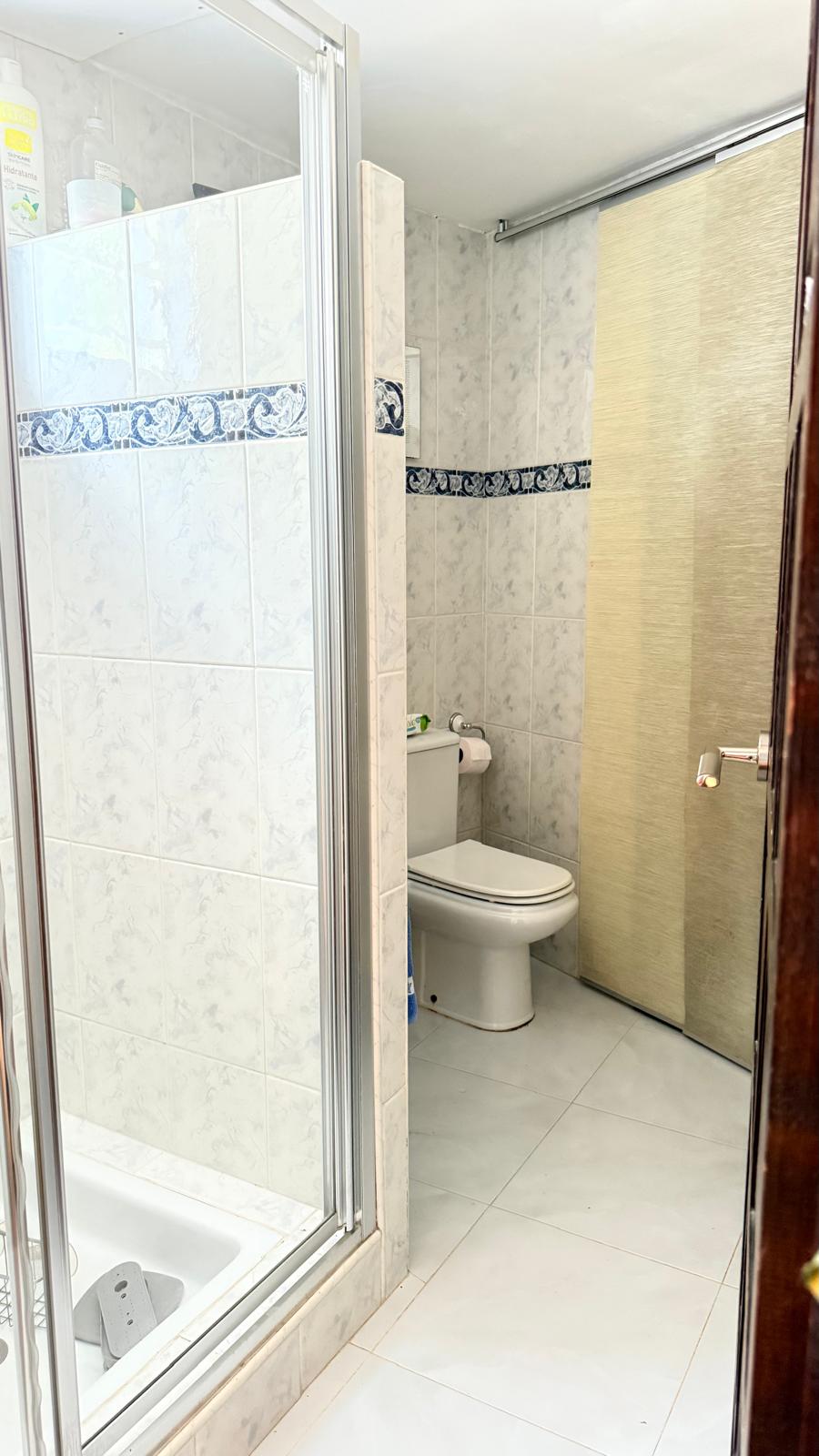 Baño con ducha acristalada, paredes alicatadas con veteado suave y cenefa decorativa azul. Cuenta con sanitarios blancos y una práctica puerta corredera.
